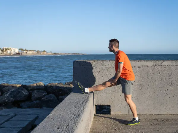 Genç sporcu deniz kenarındaki bir dalgakıranda bacaklarını esnetiyor, arka plandaki şehir manzarası ile antrenmana hazırlanıyor.