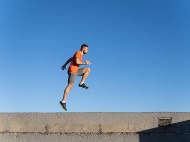 Formda sporcu dalgakıranda alçak bir duvardan atlarken arka planda açık mavi bir gökyüzü var.