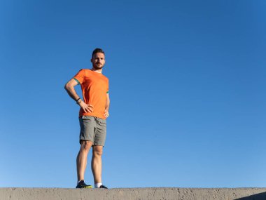 Dalgakıranın üzerinde elleri kalçasında duran genç sporcu, açık mavi gökyüzüne karşı poz veriyor.