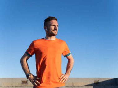 Eğitimden sonra turuncu tişört ve mavi gökyüzünün altında kablosuz kulaklık takan kendine güvenen sporcu başka yöne bakıyor.