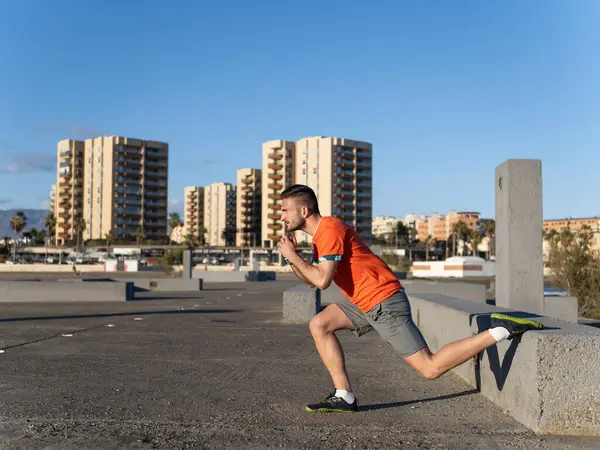 Beton bir duvarda bacak esnetme egzersizi yapan konsantre erkek sporcu. Arka planda binalar var.