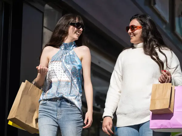 İki neşeli bayan arkadaş alışveriş torbaları taşıyor ve güneşli bir günde şehir caddesinde yürürken sohbet ediyorlar.
