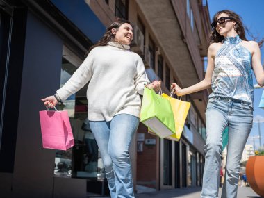 İki arkadaş alışveriş çılgınlığının tadını çıkarıyor, renkli çantalar taşıyor ve güneşli bir günde sokakta yürüyor.