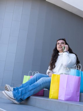 Genç bir kadın, canlı alışveriş torbalarıyla çevrili merdivenlerde oturuyor, canlı bir telefon görüşmesi yapıyor, mutluluk ve tarz saçıyor.