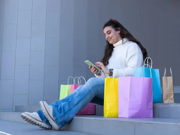 Mutlu müşteri cep telefonu uygulaması kullanıyor, online alışverişleri kontrol ediyor, alışveriş merkezindeki başarılı gününden sonra renkli alışveriş torbalarıyla merdivenlerde dinleniyor.