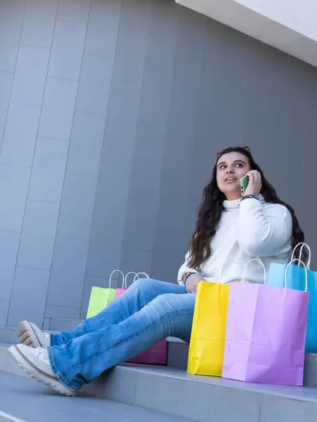 Genç bir kadın, canlı alışveriş torbalarıyla çevrili merdivenlerde oturuyor, canlı bir telefon görüşmesi yapıyor, mutluluk ve tarz saçıyor.