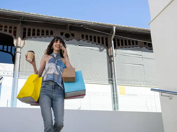 Alışveriş çılgınlığından zevk alan genç bir kadın cep telefonuyla sohbet ederken renkli poşetler ve kahve taşıyor.