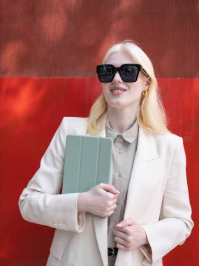 İş kadını elinde tablet, kırmızı arka planda gülümserken güneş gözlüğü takıyor.