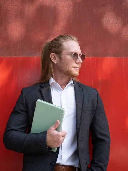 Güneş gözlüklü şık bir işadamı elinde tabletle kırmızı bir duvara yaslanırken yüzünü çeviriyor.