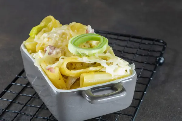 Pasta gratin with leeks and bacon, baked au gratin with grated Emmental, in a grey and white enamelled rectangular pot, placed on a black mesh trivet.