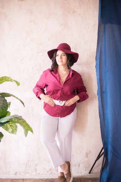 stock image A young girl stylist with dark hair smiles in a trendy burgundy color shirt and light trousers and a wide-brimmed burgundy hat in a bright studio with green living plants in pots