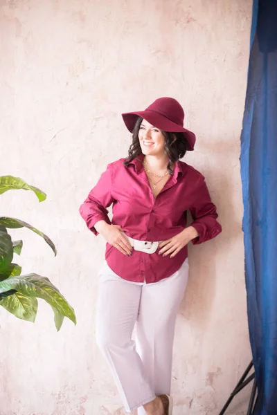 一位年轻的女造型师 黑头发 身穿时髦的勃艮第色系衬衫和浅色长裤 头戴宽边勃艮第帽 在一个明亮的工作室里 盆栽着绿色的活植物 微笑着 — 图库照片