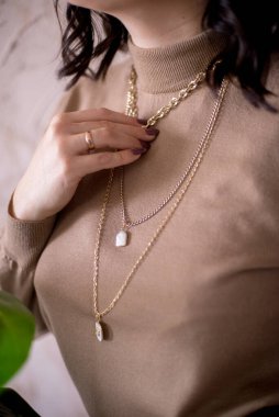 Siyah saçlı genç bir stilist Mocha golf modasında altın mücevherler içinde yeşil bitkilerle dolu bir stüdyoda gülümsüyor.