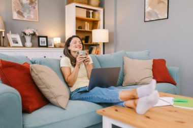 Dizüstü bilgisayarıyla kanepede oturmuş çay içen bir kadın.