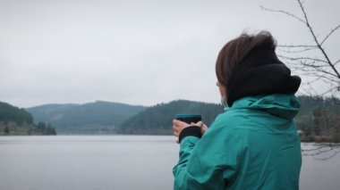 Sonbahar bulutlu bir günde elinde fincanla gezen, çay içen ve gölde dinlenen bir kadın. Aktif yaşam tarzı konsepti. Seyahat ve turizm. Sonbaharda çaydan hoşlanan bir kadın.