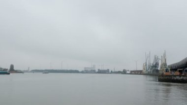 Antwerp Uluslararası Liman. Belçika, Antwerpen 'de endüstri. Sisli dumanlı bir günde arka planda vinç ve yel değirmenleriyle kanal boyunca yüzen nakliye gemileri. Yeşil enerji
