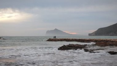 Bulutlu arka planda resimli deniz kıyısı. Büyük deniz dalgaları kayalık sahile çarpıyor ve su sıçrıyor. Güzel kayalık dağ kıyısı