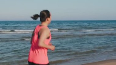 Sabah plajda koşan bir kadının sırt çekimi. Genç, zinde bir kadın deniz kıyısında koşuyor. Fitness sabahı aktivitesi. Antrenman egzersizleri. Aktif sağlıklı yaşam tarzı. Koşu ve koşu konsepti