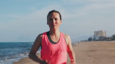 Sabahları kumlu sahil boyunca koşan kaslı vücutlu sağlıklı genç bir kadının portresi. Gülümseyen kadın plajda koşuyor, spor yapıyor. Etkin spor konsepti