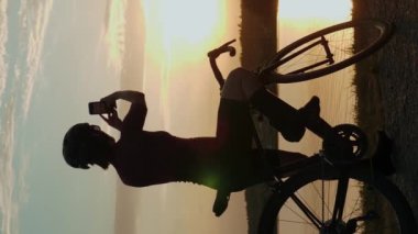 Bisikletçi bisiklete bindikten sonra gün batımının güzel fotoğraflarını çekiyor. Dikey video. Akıllı telefondan fotoğraf çeken kadın sporcu gölün kenarında durup gün batımını seyrediyor. Aktif yaşam tarzı. Boş zaman aktivitesi