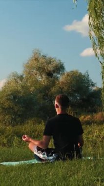 Erkek, parkta yoga minderi üzerinde meditasyon yapıyor. Dikey video. Nilüfer çiçekli halının üzerinde oturan ve yoga yapan atletik bir adam. Yogacı adam doğada dinleniyor ve temiz havanın tadını çıkarıyor. Kişisel bakım kavramı