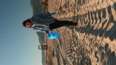 Plajda çöp toplayan koruyucu eldivenli bir kız. Dikey video. Plajda plastik temizleyen genç bir kadın. Çöp torbasında çöp toplayan bir gönüllü. Plastik kirliliği