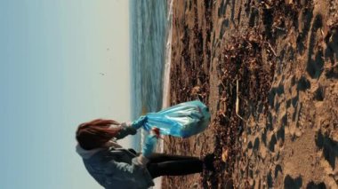 Koruyucu eldivenli genç bir kadın çöp torbasında çöp topluyor. Dikey video. Plajda plastik çöp toplayan gönüllü bir kız. Çevresel bir sorun. Yeşil Gezegen