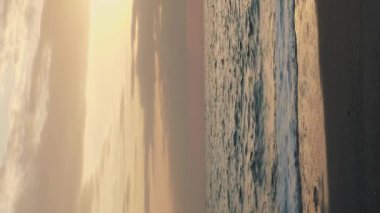 Pembe bulutlu deniz batımı. Dikey video. Güneş ufuktan batıyor. Altın gün batımında kumlu sahile ulaşan deniz dalgaları. İnanılmaz deniz doğası manzarası.