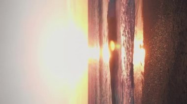 Ufukta altın deniz batımı var. Dikey video. Deniz ufkunda güneş batıyor, dalgalar yavaşça kumlu sahile ulaşıyor. Deniz Burnu