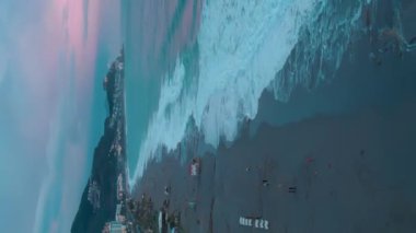 Renkli tropik plaj günbatımı. Deniz üzerinde pembe bulutlu gün batımı. Dikey video. Akşam karanlığında kumsalda duran ve ufuktaki renkli günbatımını seyreden insanlar.