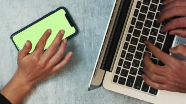 Ofiste birlikte çalışan insanlar. Dikey video. İş arkadaşları yeşil ekranlı akıllı telefon ve dizüstü bilgisayar kullanıyor, mesaj yazıyor ve çevrimiçi iletişim kuruyor. Takım çalışması. İş konsepti