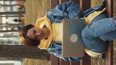 Genç serbest çalışan kadın parkta oturuyor ve dizüstü bilgisayarla çalışıyor. Dikey video. Green City Park 'ta bankta oturan ve dizüstü bilgisayar kullanan, internetten ders çalışan bir kadın. Serbest uzaktan çalışma