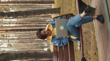 Şehir parkında bankta otururken dizüstü bilgisayar kullanan serbest çalışan bir kadın. Dikey video. Genç kız öğrenci dışarıda çalışıyor, dizüstü bilgisayar kullanıyor. Uzak çalışma
