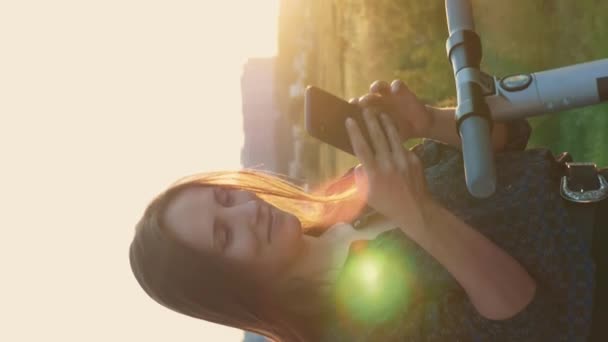 生態系輸送 シティパークで電動スクーターを使用している女性 垂直ビデオ 彼女の電動スクーターの横に立って夕日の日差しの中で女性の肖像画 生態的私的輸送を利用する者 — ストック動画