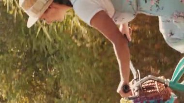 Hasır şapkalı neşeli bir kadın yazın geçmişe dönük bisiklete biniyor. Dikey video. Çiçekli elbiseli güzel esmer kadın parkta antika bir bisiklete biniyor, yaprakların arasında güneş ışığı parlıyor.