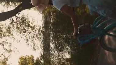 Hasır şapkalı esmer kadın nehir boyunca retro bisiklet sürüyor. Dikey video. Nehir ya da göl kenarındaki parkta sepeti ve çiçekleri olan mavi vintage bir bisiklette hareket eden güzel bir kadın.