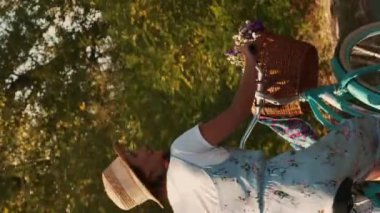 Sepeti ve çiçekleri olan eski bir bisiklete binen çekici bir kadın. Dikey video. Hasır şapkalı ve çiçekli elbiseli mutlu bir kadın parkta bisiklet sürüyor, yaz tatilinin tadını çıkarıyor.