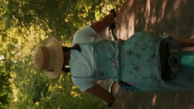 Kırsalda hasır şapkalı esmer bir kadın bisiklete biniyor. Dikey video. Elbiseli ve şapkalı genç bir kadın klasik bisiklete pedallar basıyor sepetli ve ormandaki çiçeklerle. Eğlence etkinliği