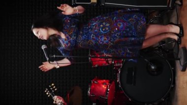 Mikrofona şarkı söyleyen mutlu, büyüleyici kadın müzisyen. Dikey video. Müzik grubu prova stüdyosunda prova yapıyor, müzik enstrümanları çalıyor ve şarkı söylüyor. Gitarist gitar çalıyor.