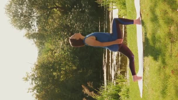 户外运动瑜伽 女人在公园里做伸展运动 垂直录像 日落时分 年轻迷人的女运动员在湖边的瑜伽垫上练习瑜伽 积极的生活方式 — 图库视频影像