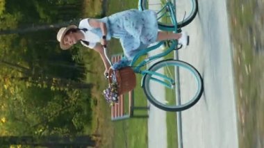 Yazın parkta geçmişe dönük bisiklet süren hasır şapkalı bir kadın. Dikey video. Genç ve çekici bir bayan, güneşli bir günde antika bir bisiklet sürüyor. Boş zaman aktivitesi. Açık hava eğlencesi