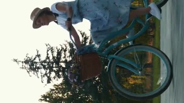 バスケットや花とヴィンテージ自転車に乗る女性サイクリスト 垂直ビデオ 夕日の街の公園で彼女のレトロな自転車でドレスやわら帽子サイクリングの若い魅力的な女性 アウトドアレジャー活動 — ストック動画