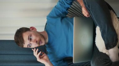 Evde dizüstü bilgisayarla çalışırken akıllı telefondan konuşan genç bir erkek. Dikey video. Ev işyerindeki rahat koltukta otururken bilgisayar başında çalışan bir adam. Şirket iş konsepti