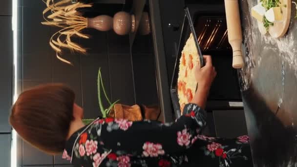 一个女人把新鲜的披萨面团放在现代厨房的烤箱里 垂直录像 女人们把意大利披萨放进电炉 准备自制美味的披萨 烹调健康食品的过程 — 图库视频影像