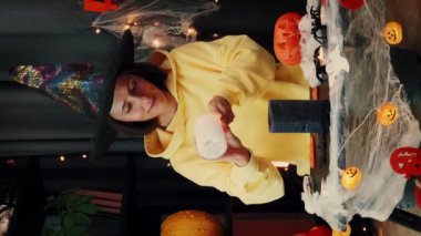 Karanlık odada oturup Cadılar Bayramı 'nı kutlayan heyecanlı kız. Portakal renkli balkabakları, iskelet ve örümceklerle Cadılar Bayramı süslemesi. Cadılar Bayramı geçmişi. Dikey video