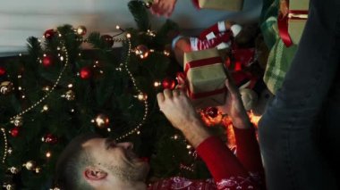Mutlu bir aile Noel 'i evde kutluyor, kırmızı kurdeleye sarılmış hediyeleri açıyor. Genç çift Noel ağacının yanında hediyelerle oturuyor ve tatilin tadını çıkarıyor. New York ruhu. Dikey video.