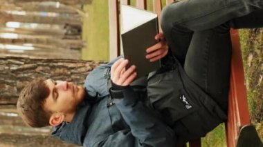 Yakışıklı sakallı adam elinde bir kitapla parkta oturuyor, sayfaları çevirip okumaya başlıyor. Sonbahar sonbahar parkında kitap okuyan genç bir erkek. Park konseptini okuyorum. Dikey video
