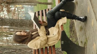 Parkta bankta oturan ve kahverengi paltolu ve atkılı kitap okuyan hoş bir kız. Yakışıklı esmer kadın sonbahar parkında kitap okuyor. Okuma konsepti. Dikey video