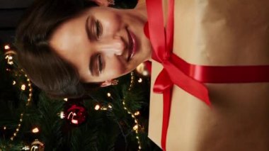 Noel hediyesiyle hediye kutusunu sallayan mutlu genç bir kadın Noel ağacının yanında oturuyor. Kadın Noel hediyesini kırmızı kurdeleyle sarmış. Yılbaşı ve Noel arifesi kutlaması. Dikey video