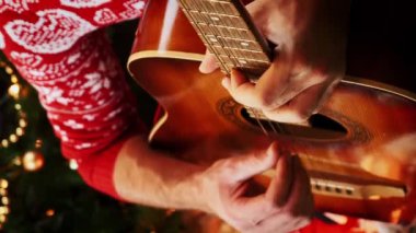 Şenlikli kırmızı kazaklı bir adam Noel arifesini kutlamak için Noel şarkılarını gitarla çalarak süslenmiş yılbaşı ağacının yanında oturuyor. Mutlu Noeller, mutlu yıllar. Dikey video